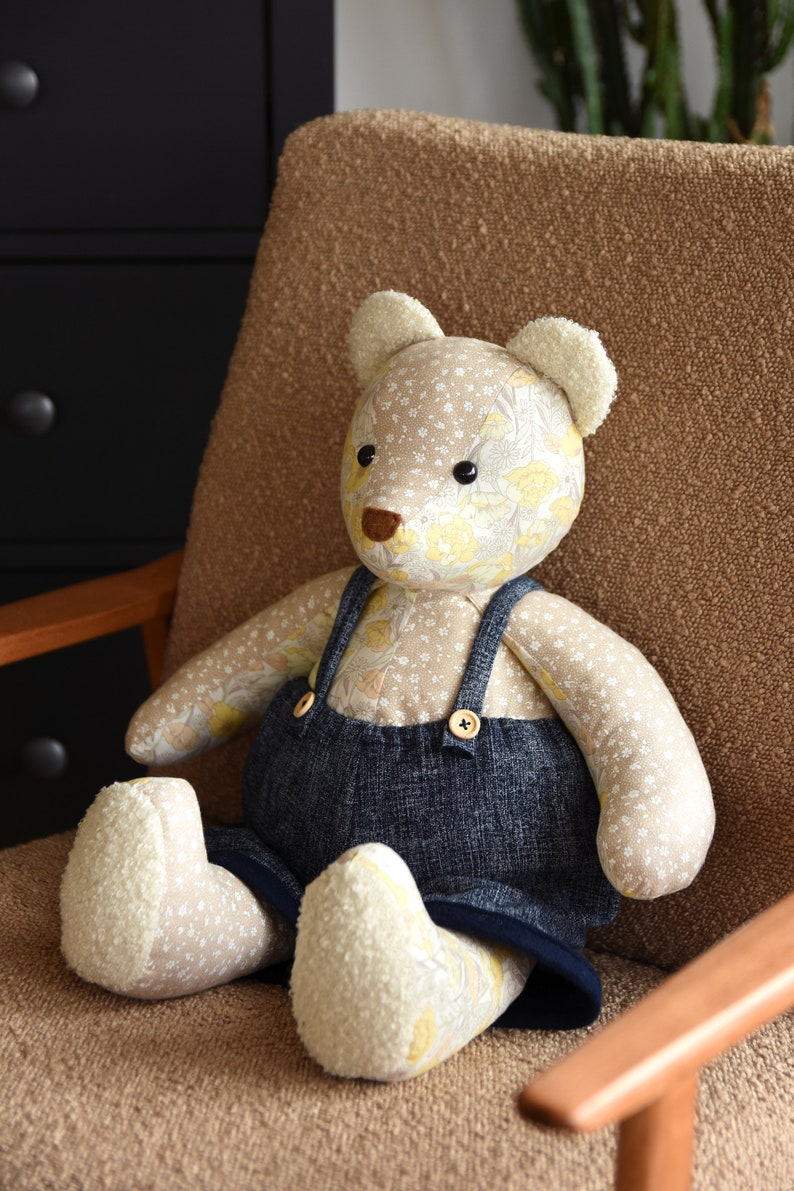 Puede incluir: Un oso de peluche hecho a mano con un cuerpo estampado floral y un peto vaquero. El oso tiene orejas y pies color crema, una nariz marr&oacute;n y ojos de bot&oacute;n negros. Est&aacute; sentado en una silla marr&oacute;n.