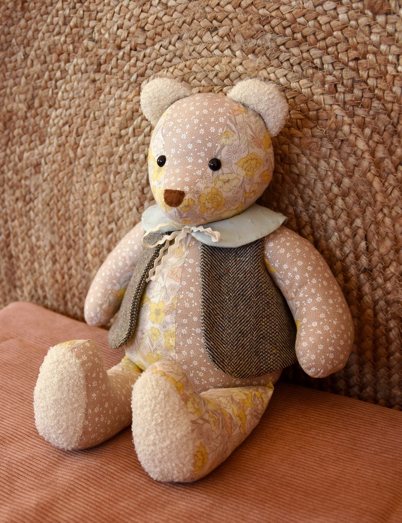 May include: A plush teddy bear with a floral pattern and a brown tweed vest. The bear is sitting on a brown corduroy surface.