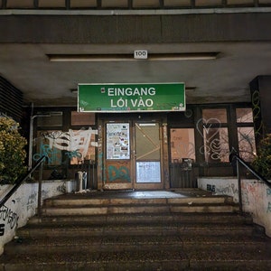 May include: Exterior shot of a building entrance at night. A green sign above the door reads "Eingang Lôi Vao". The entrance has a set of stairs leading up to a wooden door. Graffiti is visible on the walls and doors.
