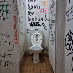 May include: A public restroom with graffiti-covered walls and a toilet. The walls are covered in various writings and drawings in multiple colors. The text "Lets lynch the landlord" is visible on the wall above the toilet.