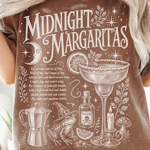 May include: A brown short-sleeve t-shirt featuring white graphic illustrations of a margarita glass, a frog wearing a witch hat, a poison bottle, and a coffee pot. The text reads Midnight Margaritas and includes a poem about a witch's brew.
