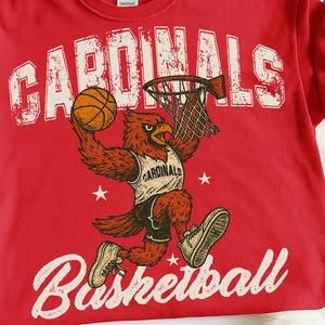 May include: Red t-shirt with a distressed graphic of a cardinal bird playing basketball. The bird is wearing a jersey that says "Cardinals." The word "Cardinals" is printed above the bird, and "Basketball" is printed below.