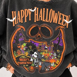 May include: A charcoal gray crewneck sweatshirt featuring a large graphic print with the text Happy Halloween in orange. The design includes white bats, a pumpkin-shaped border, and a scene of Mickey Mouse in a pumpkin patch.