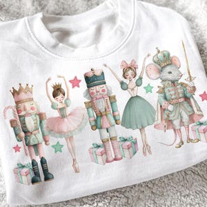 Peut inclure: Sweat-shirt blanc avec un motif sur le thème de Casse-Noisette. L'illustration comprend un casse-noisette, des danseuses et un roi des souris, le tout dans des couleurs pastel. Des boîtes cadeaux et des étoiles roses et vertes complètent le design.