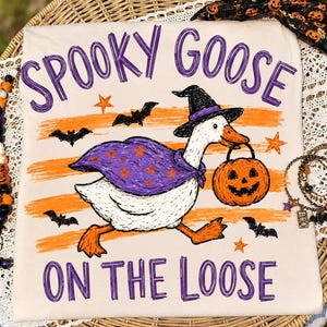 May include: A cream-colored t-shirt featuring a graphic of a white goose wearing a purple cape with orange stars and a black witch hat. The goose carries an orange jack-o'-lantern bucket. The text reads SPOOKY GOOSE ON THE LOOSE with bats.