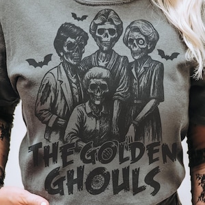 Puede incluir: Camiseta gris con un gr&aacute;fico negro de cuatro figuras esquel&eacute;ticas y murci&eacute;lagos. El texto "THE GOLDEN GHOULS" est&aacute; impreso debajo de las figuras en una fuente desgastada. La camiseta tiene mangas cortas y cuello redondo.