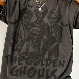 Puede incluir: Camiseta gris oscuro con un gr&aacute;fico de cuatro figuras esquel&eacute;ticas y el texto "THE GOLDEN GHOULS". El dise&ntilde;o incluye murci&eacute;lagos. La camiseta tiene un aspecto vintage y desgastado.