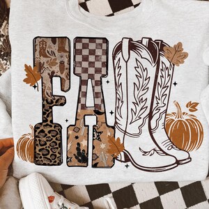 K&ouml;nnte beinhalten: Ein hellgraues Sweatshirt mit dem Wort "FA" in einer dekorativen Schriftart. Der Buchstabe "F" hat ein Leopardenmuster und der Buchstabe "A" hat ein Schachbrettmuster. Das Design umfasst Cowboy-Stiefel, K&uuml;rbisse und Herbstbl&auml;tter.