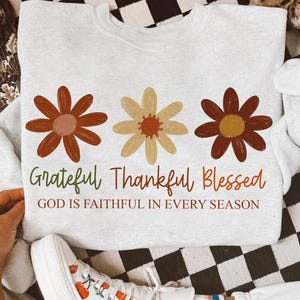 May include: A light gray sweatshirt featuring three stylized flowers in shades of brown and yellow. The words "Grateful Thankful Blessed" are printed below the flowers, with the phrase "GOD IS FAITHFUL IN EVERY SEASON" underneath.