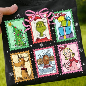 May include: Black t-shirt featuring a colorful Christmas-themed design. The design includes images of a Christmas tree, gifts, the Grinch, Max the dog, and Cindy Lou Who, all framed as postage stamps. A pink bow and snowflakes complete the festive look.
