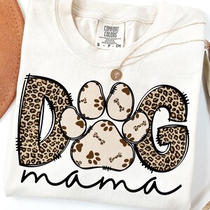 Pu&ograve; includere: Una maglietta bianca con un design a stampa leopardata marrone e nera che dice "Dog mama". Il "Dog" &egrave; composto da un'impronta di zampa e le lettere "D" e "G" sono in stampa leopardata.