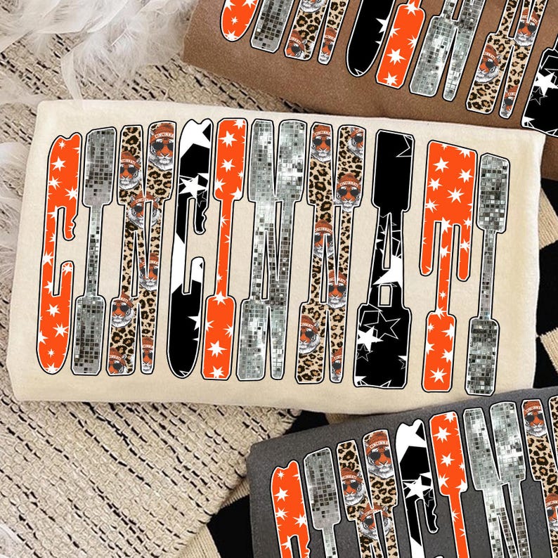 Puede incluir: Camiseta color crema con la palabra "CLENCH" en un dise&ntilde;o &uacute;nico. Las letras presentan patrones naranja, plateado, negro y de leopardo, con detalles de estrellas y un tigre con gafas de sol. Se ven varias otras camisetas con el mismo dise&ntilde;o.