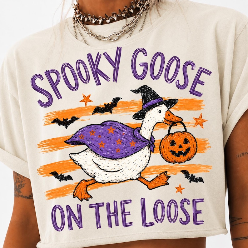 May include: A beige t-shirt featuring a graphic of a white goose wearing a black witch hat and a purple cape with orange stars. The goose carries an orange jack-o'-lantern bucket. The text reads SPOOKY GOOSE ON THE LOOSE with orange stripes and bats.