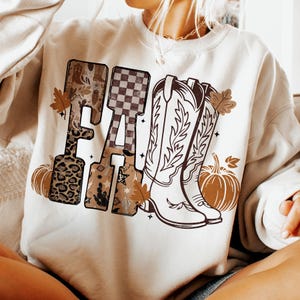 K&ouml;nnte beinhalten: Cremefarbenes Sweatshirt mit dem Wort "FALL" in dekorativer Schrift. Das "F" hat einen Leopardenmuster, das "A" hat ein Schachbrettmuster und das "L" hat ein W&uuml;sten-Design. Cowboy-Stiefel, K&uuml;rbisse und Herbstbl&auml;tter vervollst&auml;ndigen das Design.