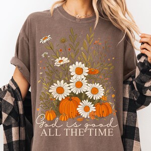 May include: A brown t-shirt with a floral design including white daisies, orange pumpkins, and green leaves. The text "God is good ALL THE TIME" is printed in a script font. The shirt has a relaxed fit.