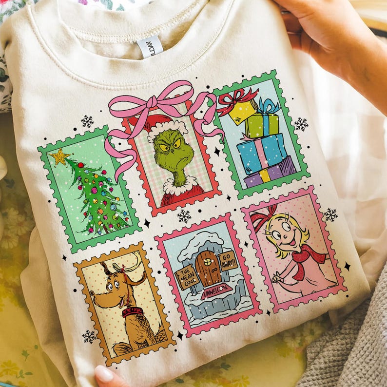 May include: Cream-colored sweatshirt featuring a colorful graphic of Christmas-themed postage stamps. The stamps depict a Christmas tree, the Grinch, wrapped gifts, Max the dog, Cindy Lou Who, and a snowy scene. A pink bow is at the top.