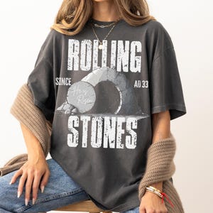 May include: Dark gray t-shirt with the text "ROLLING STONES" and "SINCE AD 33". The graphic features a stone archway and the words "He is Risen" on a stone. The shirt is a relaxed fit.