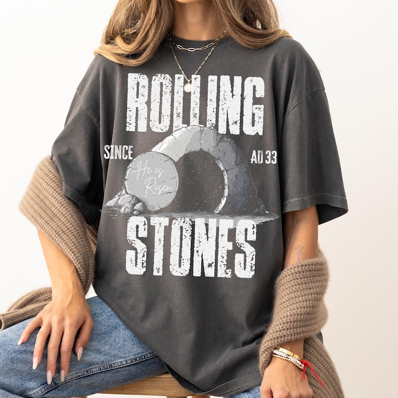 May include: Dark gray t-shirt with the text "ROLLING STONES" and "SINCE AD 33". The graphic features a stone archway and the words "He is Risen" on a stone. The shirt is a relaxed fit.