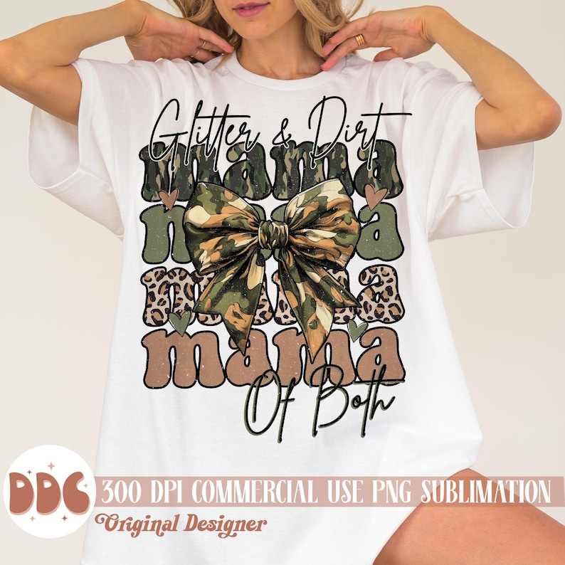 以下が含まれることがあります： 白地に「Glitter & Dirt Mama Of Both」という茶色と黒の文字が書かれたグラフィックデザインのTシャツ。グラフィックには、茶色のヒョウ柄のアクセントが付いた迷彩柄のリボンも描かれている。