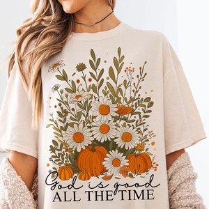 May include: A cream-coloured t-shirt with a floral design. The design includes daisies, pumpkins, and foliage in orange, green, and white. The text "God is good all the time" is written in a script font at the bottom of the design.