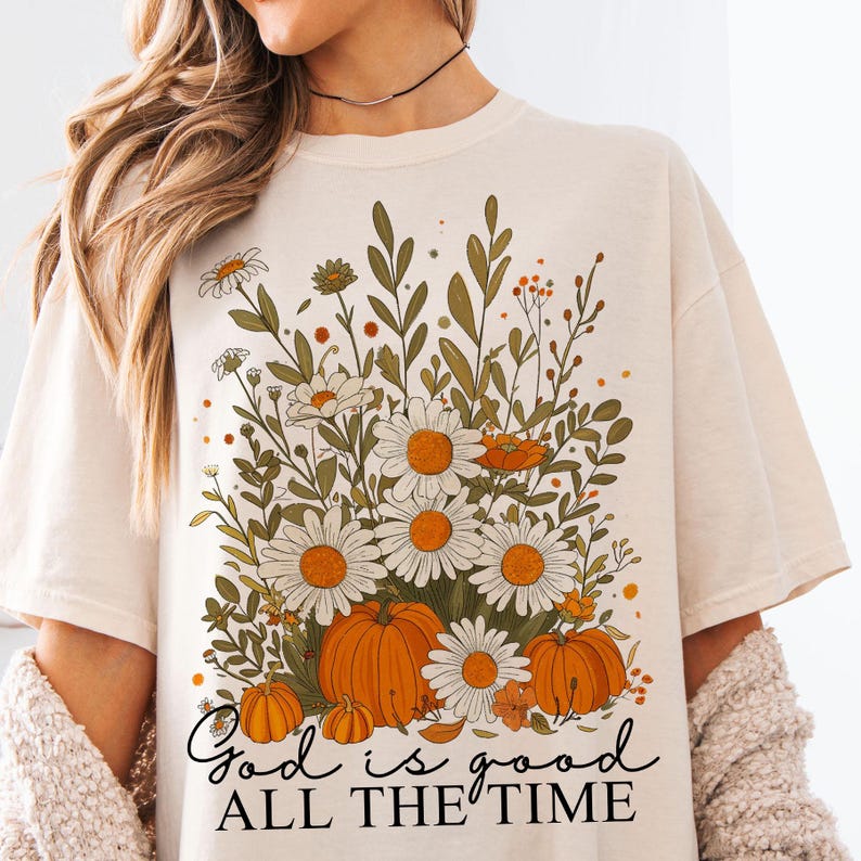 May include: A cream-coloured t-shirt with a floral design. The design includes daisies, pumpkins, and foliage in orange, green, and white. The text "God is good all the time" is written in a script font at the bottom of the design.