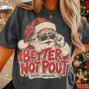 May include: A charcoal gray t-shirt featuring a graphic of Santa Claus wearing sunglasses and a red hat. The text reads "BETTER NOT POUT" in large red letters, with "SANTA'S WATCHING" printed in smaller black text underneath the main graphic.