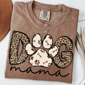 Pu&ograve; includere: Una maglietta marrone con una grafica della parola "DOG" in stampa leopardata con un'impronta di zampa al centro. La parola "mama" &egrave; scritta sotto la grafica.