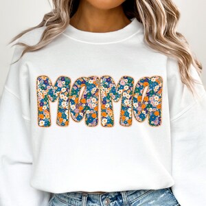 May include: White sweatshirt with the word "Mama" in large, colorful letters. The letters are covered in a floral pattern with orange, blue, and pink flowers.