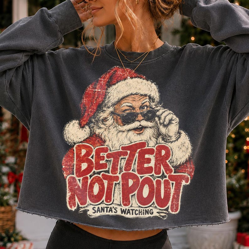 May include: A charcoal gray long-sleeve cropped sweatshirt featuring a graphic of Santa Claus wearing sunglasses. The text reads "BETTER NOT POUT" in large red distressed letters, with "SANTA'S WATCHING" printed in smaller white letters below.
