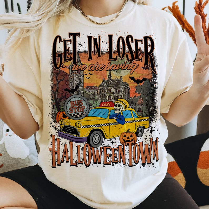 May include: Cream-colored t-shirt with a Halloween-themed graphic. The design features a yellow taxi with a skeleton driver, pumpkins, and the text "GET IN LOSER we are saving HALLOWEENTOWN".
