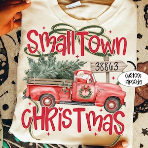 May include: Cream-colored t-shirt with a festive Christmas design. The design features a red pickup truck carrying a Christmas tree, Santa Claus, and the words "Small Town Christmas" in red. A custom zipcode is also included.