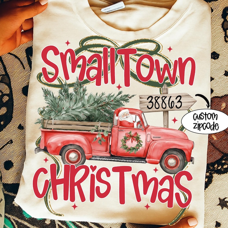 May include: Cream-colored t-shirt with a festive Christmas design. The design features a red pickup truck carrying a Christmas tree, Santa Claus, and the words "Small Town Christmas" in red. A custom zipcode is also included.