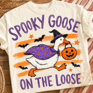 May include: A cream-colored t-shirt featuring a graphic of a white goose wearing a purple cape and a black witch hat. The goose carries an orange jack-o'-lantern. The text reads "SPOOKY GOOSE ON THE LOOSE" in purple, surrounded by orange stripes, black bats, and orange stars.