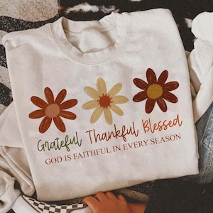 May include: Cream-colored sweatshirt featuring three stylized flowers in shades of brown and yellow. The words "Grateful Thankful Blessed" are printed below the flowers, with the phrase "GOD IS FAITHFUL IN EVERY SEASON" underneath.