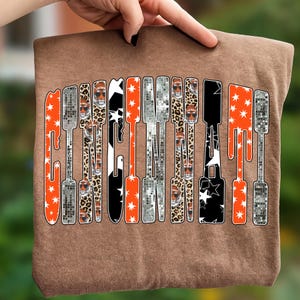 Puede incluir: Una camiseta marr&oacute;n con la palabra "CINCINNATI" impresa verticalmente en un dise&ntilde;o &uacute;nico. Las letras est&aacute;n rellenas de varios patrones, incluyendo naranja con estrellas blancas, estampado de leopardo y negro con estrellas blancas. La camiseta est&aacute; sujeta por una mano.