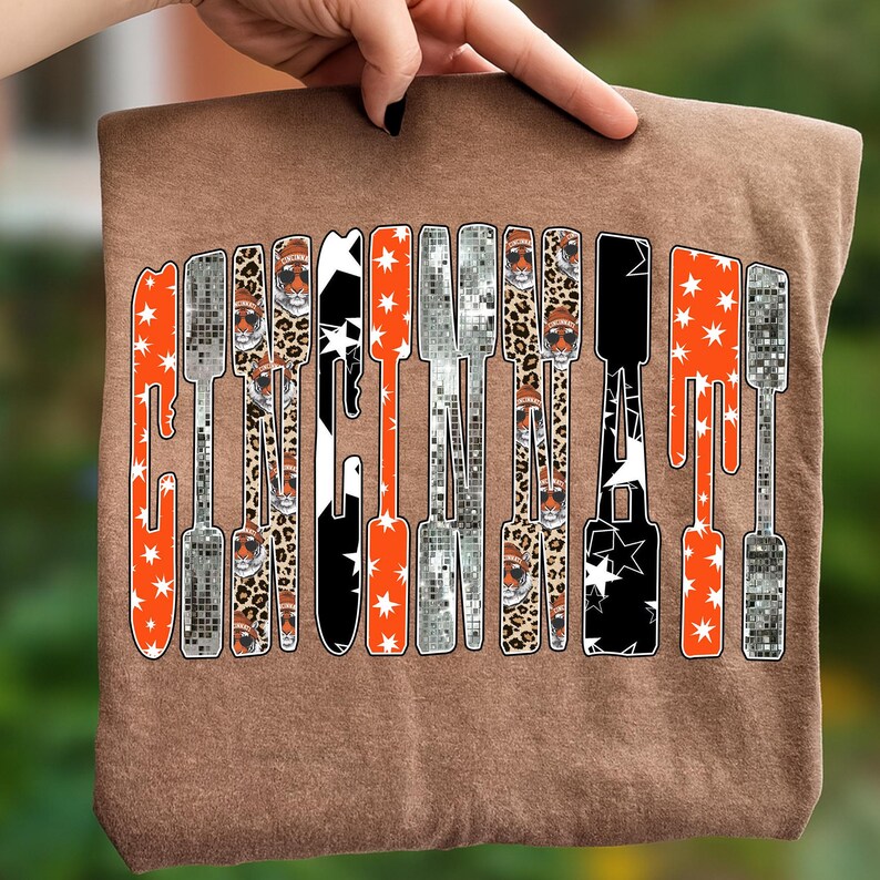Puede incluir: Una camiseta marr&oacute;n con la palabra "CINCINNATI" impresa verticalmente en un dise&ntilde;o &uacute;nico. Las letras est&aacute;n rellenas de varios patrones, incluyendo naranja con estrellas blancas, estampado de leopardo y negro con estrellas blancas. La camiseta est&aacute; sujeta por una mano.