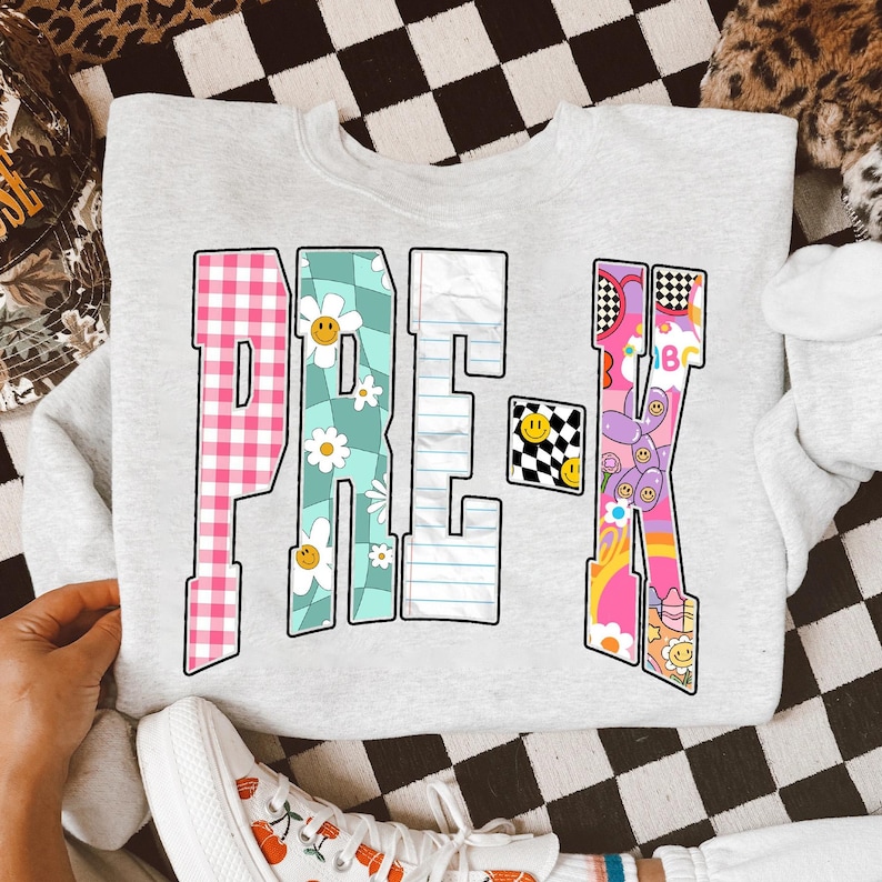 Op de afbeelding: Een lichtgrijze sweatshirt met het woord "PRE-K" in kleurrijke, patroonletters. De letters bevatten ontwerpen zoals roze ruitjes, madeliefjes, gelinieerd papier en een dambordpatroon. Een kleine smiley is ook inbegrepen.