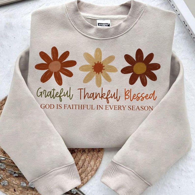 May include: Beige sweatshirt with three flower graphics in shades of brown and yellow. The text "Grateful Thankful Blessed" and "God is faithful in every season" are printed below the flowers.