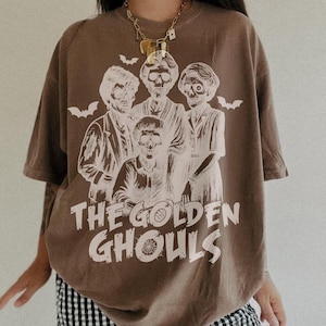 Puede incluir: Camiseta marr&oacute;n con un dise&ntilde;o blanco de cuatro figuras esquel&eacute;ticas, murci&eacute;lagos y el texto "THE GOLDEN GHOULS". La camiseta tiene mangas cortas y un corte holgado, ideal para un look casual.