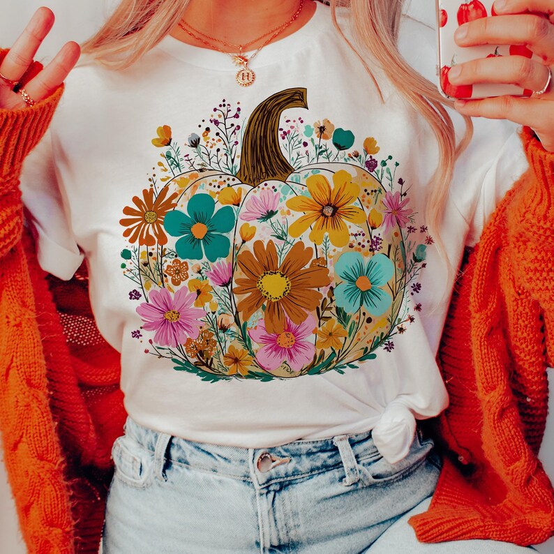 May include: White t-shirt featuring a floral pumpkin design. The pumpkin is filled with colorful flowers in shades of teal, yellow, pink, and brown. The shirt has short sleeves and a tied knot at the waist, perfect for fall.