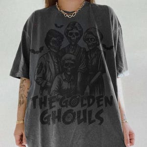 Puede incluir: Camiseta gris oscuro con un gr&aacute;fico de cuatro figuras esquel&eacute;ticas y el texto "THE GOLDEN GHOULS" en negro. El dise&ntilde;o incluye siluetas de murci&eacute;lagos. La camiseta tiene mangas cortas y un corte holgado.