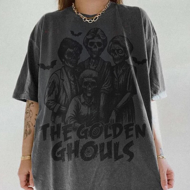 Puede incluir: Camiseta gris oscuro con un gr&aacute;fico de cuatro figuras esquel&eacute;ticas y el texto "THE GOLDEN GHOULS" en negro. El dise&ntilde;o incluye siluetas de murci&eacute;lagos. La camiseta tiene mangas cortas y un corte holgado.