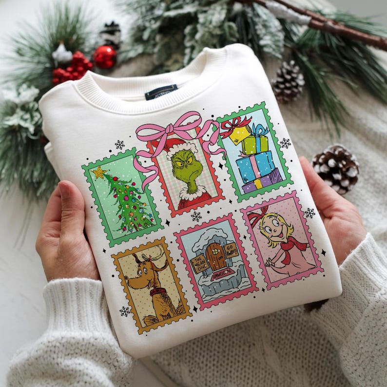 May include: Cream-colored sweatshirt featuring a colorful graphic of postage stamps with characters and scenes from a holiday story. The stamps include a Christmas tree, gifts, and a green character with a Santa hat.