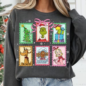 May include: Dark gray sweatshirt featuring a colorful graphic of Christmas-themed postage stamps. The stamps depict a Christmas tree, the Grinch, wrapped gifts, Max the dog, a sign that says "The Mean One", and Cindy Lou Who.