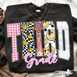 Op de afbeelding: Zwart sweatshirt met de woorden "THIRD Grade" in een kleurrijk, retro ontwerp. De "T" is roze ruitjes, de "H" en "I" hebben smiley gezichten en dambordpatronen, en de "R" heeft bloemen. De "D" is gelijnd papier.