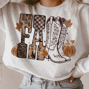 K&ouml;nnte beinhalten: Ein hellgraues Sweatshirt mit einem herbstlichen Motiv. Das Wort "FALL" wird mit jedem Buchstaben in einem anderen Muster angezeigt, darunter Leopardenmuster, Schachbrettmuster und ein s&uuml;dwestliches Design. Cowboy-Stiefel und K&uuml;rbisse vervollst&auml;ndigen das Design.