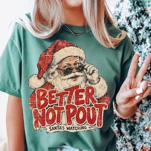 May include: A forest green short-sleeve t-shirt featuring a graphic of Santa Claus wearing sunglasses. The text reads "BETTER NOT POUT" in large red letters, with "SANTA'S WATCHING" printed below in smaller black text. The design has a distressed finish.
