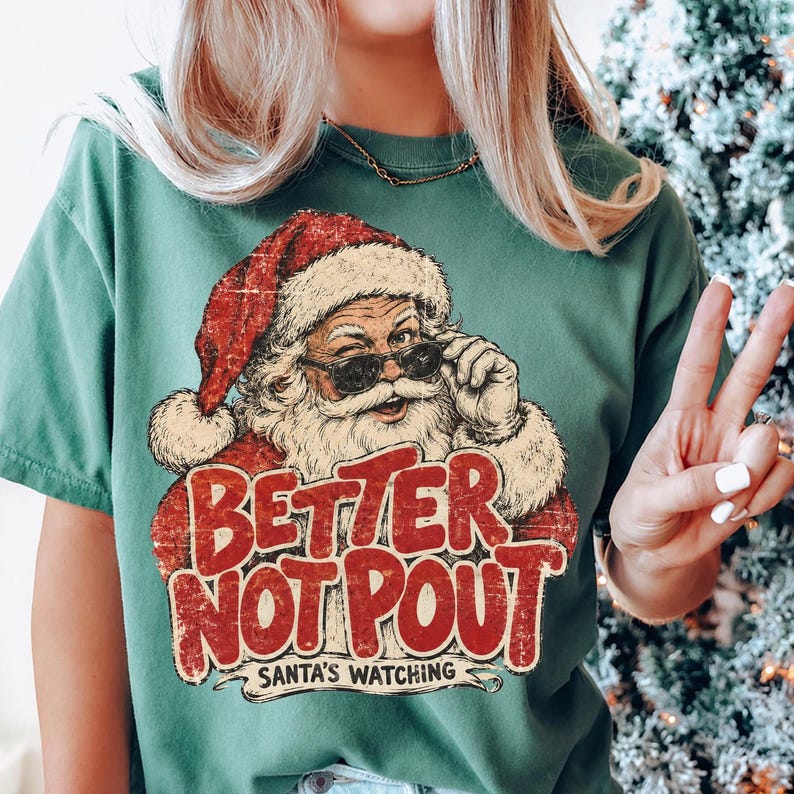 May include: A forest green short-sleeve t-shirt featuring a graphic of Santa Claus wearing sunglasses. The text reads "BETTER NOT POUT" in large red letters, with "SANTA'S WATCHING" printed below in smaller black text. The design has a distressed finish.