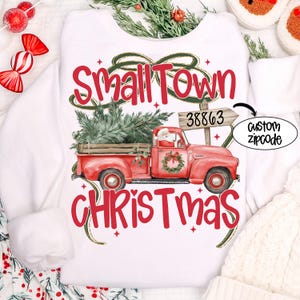 May include: White sweatshirt with a festive design. Features a red truck carrying a Christmas tree, with the words "Small Town Christmas" in red. Includes a custom zipcode option.