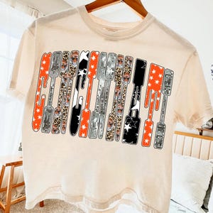 Puede incluir: Camiseta color crema con la palabra "Guitar" en un dise&ntilde;o estilizado. Las letras est&aacute;n compuestas por varios patrones, incluyendo naranja con estrellas blancas, estampado de leopardo, negro con estrellas blancas y patrones de bola de discoteca plateada.